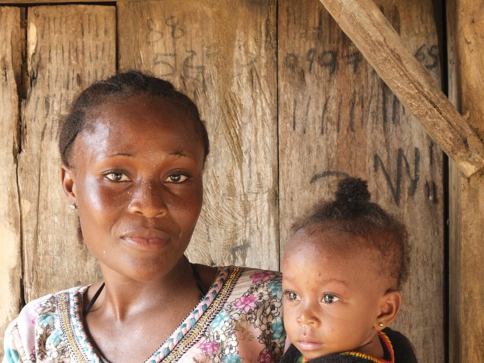 FGM Junge afrikanische Frau aus Sierra Leone, die sich gegen weibliche Gentialverstümmelung ausspricht, mit ihrem Baby auf dem Arm vor einer Holztür