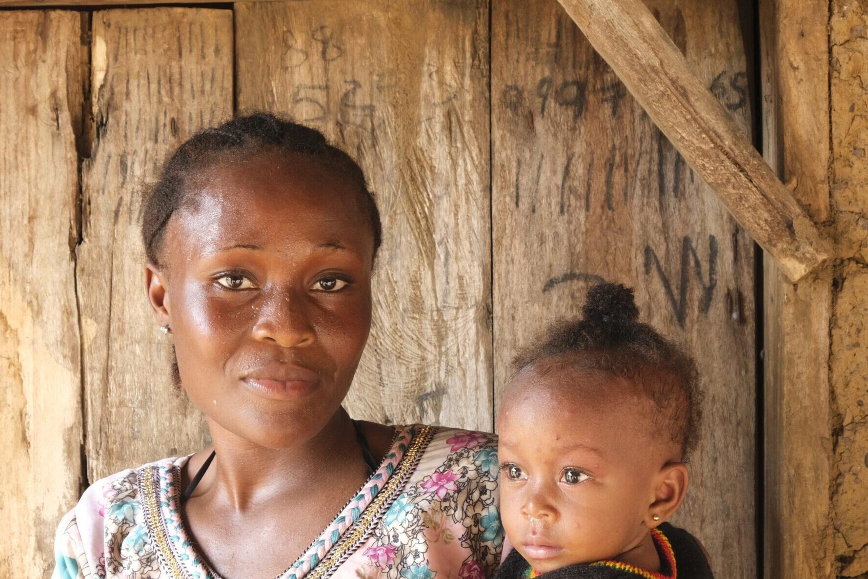 FGM Junge afrikanische Frau aus Sierra Leone, die sich gegen weibliche Gentialverstümmelung ausspricht, mit ihrem Baby auf dem Arm vor einer Holztür