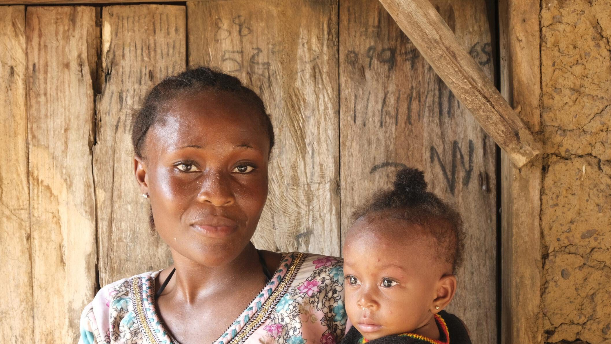 FGM Junge afrikanische Frau aus Sierra Leone, die sich gegen weibliche Gentialverstümmelung ausspricht, mit ihrem Baby auf dem Arm vor einer Holztür