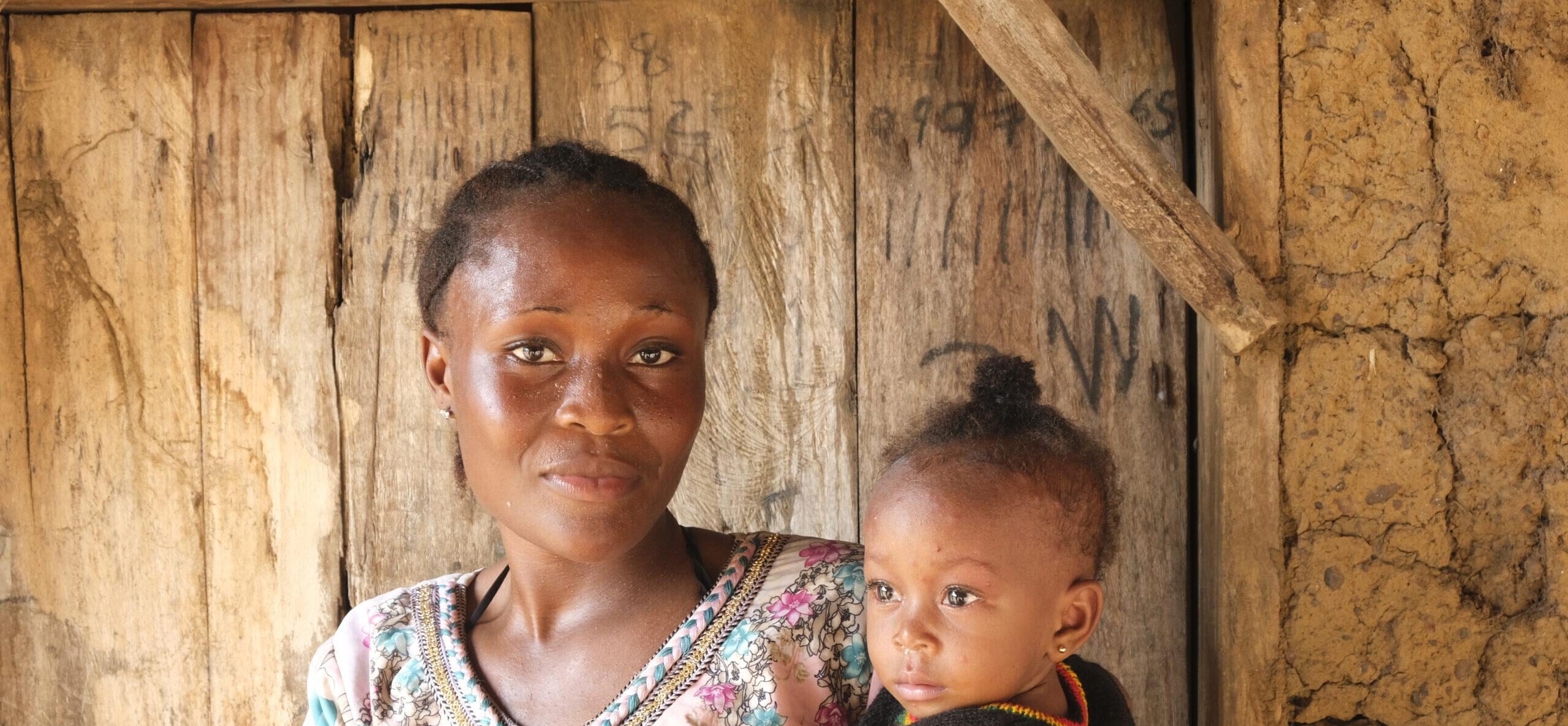 FGM Junge afrikanische Frau aus Sierra Leone, die sich gegen weibliche Gentialverstümmelung ausspricht, mit ihrem Baby auf dem Arm vor einer Holztür