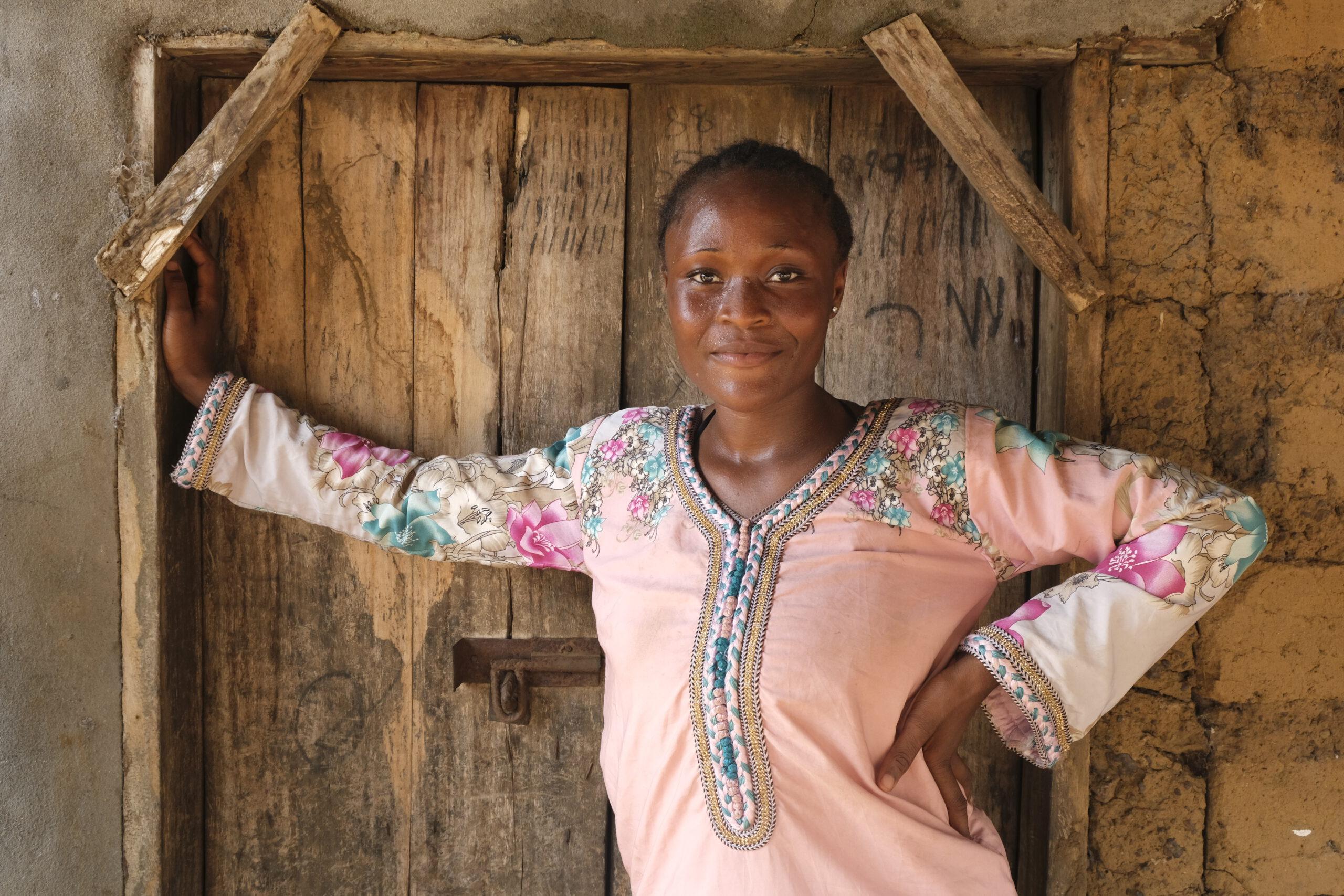 Junge sierra leonische Gegnerin von FGM stützt selbstbewusst und lächelnd einen Arm in den Rahmen einer Holztür, den anderen in ihre Hüfte